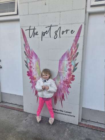 A smiling young girl stands in front of colorful angel wings painted on a wall, appearing to have wings herself. The wall says “the pet store.”