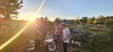 Cuatro personas se reúnen en torno a una mesa al aire libre, preparando la comida al atardecer en un campo rústico cubierto de hierba. La luz dorada crea un ambiente cálido y relajado.