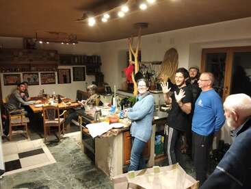 Eine lebhafte Küchenszene mit acht Personen; einige unterhalten sich um einen Holztisch herum, andere stehen an der Kücheninsel, lächeln und posieren für die Kamera.