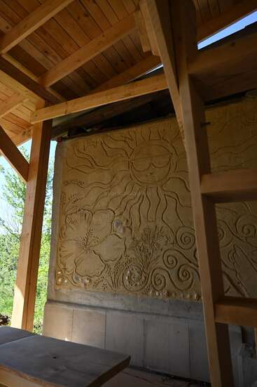 Cette image montre une charpente en bois, un mur sculpté représentant un soleil avec des rayons, des fleurs et des volutes, sous la lumière naturelle, avec une échelle en bois visible.