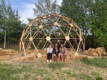 Trois personnes sont assises côte à côte sur des bottes de paille devant une grande structure géodésique en bois, entourées d'arbres et d'autres bottes de paille dans un espace vert.