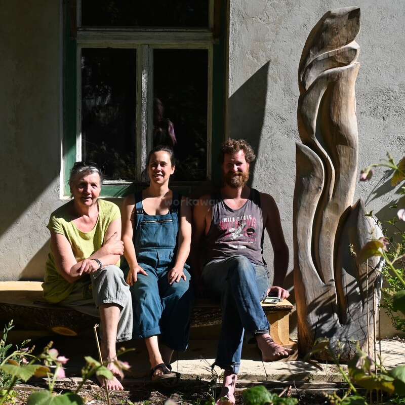 Trois personnes sont assises sur un banc devant un bâtiment rustique, le sourire aux lèvres, avec une grande sculpture abstraite en bois à leurs côtés, tandis que la lumière du soleil projette des ombres marquées tout autour.