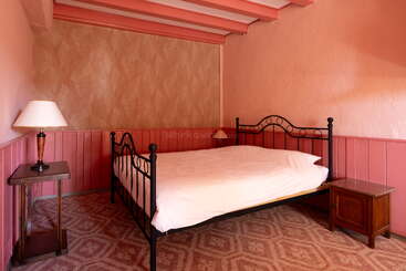 Dieses Bild zeigt ein kleines, gemütliches Schlafzimmer mit rosa Wänden und Decke, einem Bettgestell aus Eisen, weißer Bettwäsche, zwei Nachttischen aus Holz und einer Lampe.