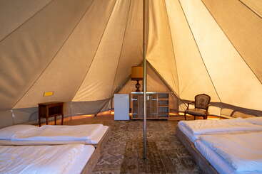 Dieses Bild zeigt das Innere eines geräumigen Glamping-Zeltes mit zwei Betten, einem Kühlschrank, einem Schreibtisch, einer Lampe, einem Sessel und einer Stange in der Mitte. Gemütliche Atmosphäre.