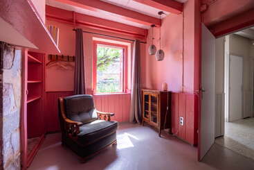 Dieses gemütliche Zimmer hat rosa Wände und Deckenbalken, einen großen Sessel, einen Holzschrank, ein Fenster mit grauen Vorhängen, einen offenen Schrank und hängende Pendelleuchten.