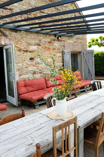 Eine rustikale Terrasse mit einer Steinmauer, einem Holztisch, verschiedenen Stühlen, einem roten Sofa mit Kissen, einem Blumengesteck, offenen Fensterläden und einer Pergola mit Lattenrost darüber.