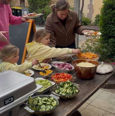 Eine Familie versammelt sich um einen Buffettisch im Freien, bedient sich aus Schalen mit frischem Gemüse, Pommes, Käse und Belägen und stellt gemeinsam im Garten ihre ganz persönlichen Gerichte zusammen.