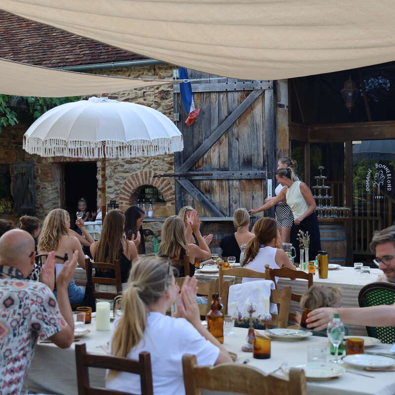 Eine Gruppe Leute genießt ein Essen im Freien an langen Tischen unter Sonnenschirmen, mit rustikaler Einrichtung, Holzstühlen und einem alten Steingebäude im Hintergrund.