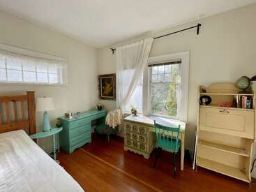 This cozy bedroom features vintage furniture, a wooden bed, turquoise desk, and chair, sheer curtains, a bookcase, soft lighting, and a large window with garden views.