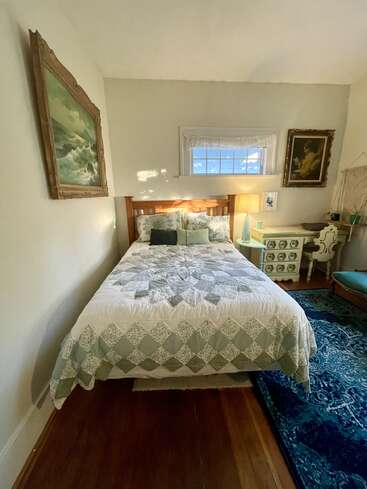 A cozy bedroom with a wooden bed, patterned quilt, blue rug, two framed paintings, window, green desk, lamp, and decorative pillows, creating a charming atmosphere.