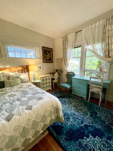 This cozy bedroom features a patterned quilt, vintage-style furniture, blue accents, large windows with sheer curtains, a decorative rug, and soft, warm lighting throughout.