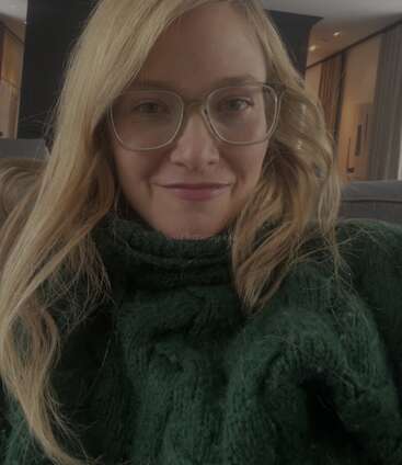 A woman with long blonde hair and glasses smiles gently. She wears a cozy, thick green sweater and sits indoors, with soft lighting in the background.