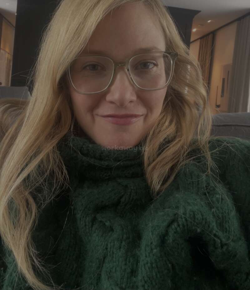 A woman with long blonde hair and glasses smiles gently. She wears a cozy, thick green sweater and sits indoors, with soft lighting in the background.