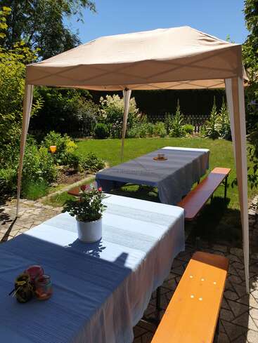 A sunlit garden setup with two long tables covered in blue cloths, wooden benches, a beige canopy, potted flower, candles, and lush green surroundings.