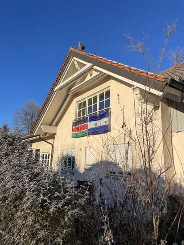A house with a steep roof, two flags hanging below the window, frosted bushes in the foreground, and a clear blue sky above.