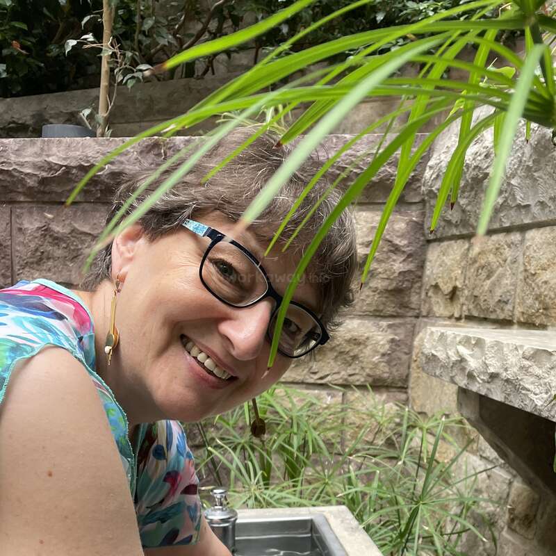 A smiling woman with glasses leans over an outdoor sink, surrounded by lush green plants and stone walls. Vibrant colors and natural light create a cheerful atmosphere.