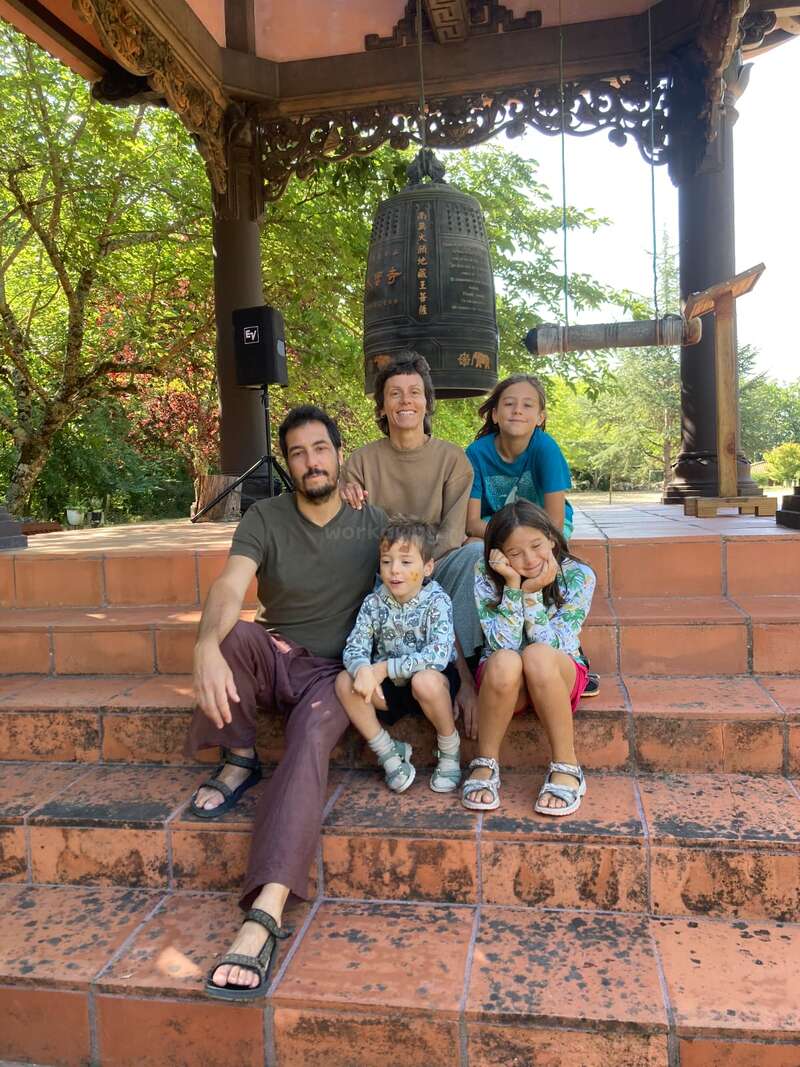 Une famille de cinq personnes est assise sur des marches en terre cuite, sous une grande cloche richement décorée, dans un cadre paisible en plein air, entourée de verdure et d'un temple à l'architecture en bois, et sourit joyeusement.