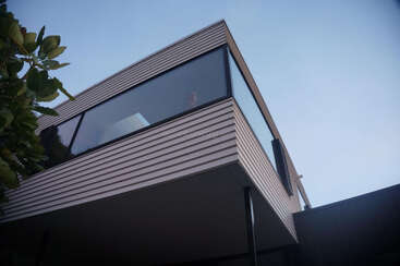 Esta imagen muestra una casa moderna con grandes ventanas de cristal y revestimiento horizontal. La perspectiva es ascendente, mostrando líneas limpias contra un cielo azul despejado.