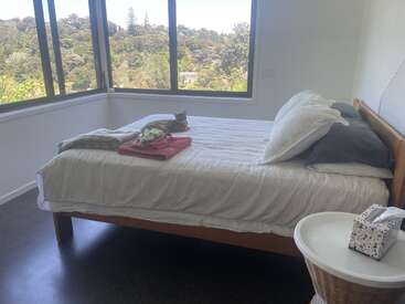 Un dormitorio luminoso con grandes ventanales con vistas a los árboles. Una cama con ropa de cama a rayas sostiene toallas dobladas y un gato. Cerca hay una mesa redonda con pañuelos.