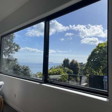 Una habitación luminosa con un gran ventanal ofrece una impresionante vista del cielo azul, las nubes esponjosas, el océano y los frondosos árboles verdes del exterior, creando tranquilidad.