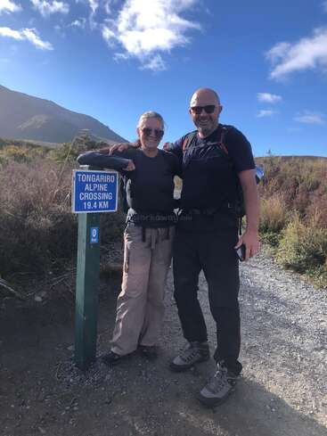 A smiling couple wearing hiking gear stands beside a "Tongariro Alpine Crossing 19.4 KM" signpost, with mountains and blue sky in the scenic background.