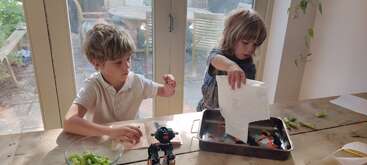 Zwei Kinder sitzen an einem Holztisch neben Glastüren. Eines spielt mit Sellerie, das andere hantiert mit Papier über einem Tablett mit LEGO Steinen, ein Roboter steht daneben.