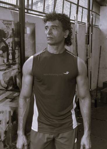 Ein muskulöser Mann mit einem ärmellosen Hemd steht in einem Fitnessstudio und schaut nach rechts. Hinter ihm sind Trainingsgeräte und Gewichte zu sehen. Das Bild ist monochrom.