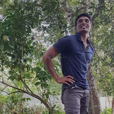 The image depicts a man standing in front of a tree, with his hand on his hip, wearing a blue shirt and grey pants, in a natural setting.