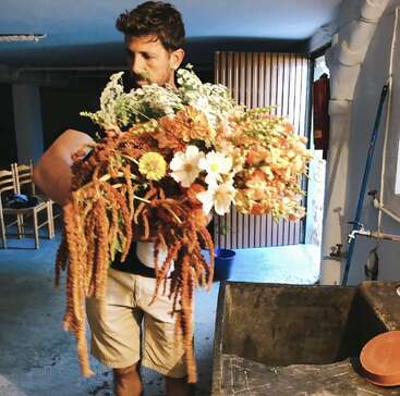 Ein Mann steht drinnen und hält einen großen, üppigen Strauß aus bunten Blumen und Grünpflanzen in der Hand; er trägt helle Shorts, hinter ihm sind Stühle und ein Waschbecken zu sehen.