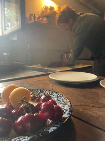 Auf einem Holztisch steht eine farbenfrohe Schale mit Kirschen und Aprikosen, während im Hintergrund eine Person in der sanft beleuchteten, gemütlichen Küche das Essen zubereitet.
