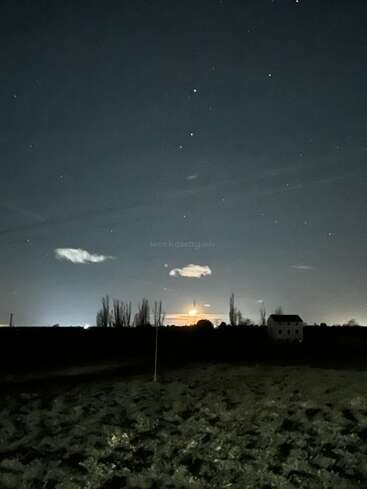 Eine friedliche Nachtlandschaft mit Sternenhimmel, einem leuchtenden Mond am Horizont, sich abzeichnenden Bäumen, einem Haus in der Ferne, Feldern und vereinzelten Wolken.