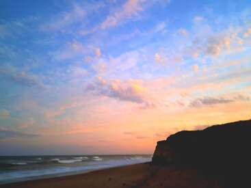 Uma praia deslumbrante ao pôr do sol apresenta nuvens coloridas em tons pastéis e uma silhueta dramática de um penhasco contra um céu brilhante com ondas suaves rolando na costa arenosa.