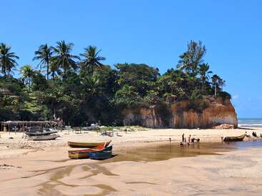Uma bela praia tropical com areia dourada, barcos coloridos, palmeiras, vegetação verde exuberante, penhasco, céu azul claro e pessoas aproveitando a água e o sol.