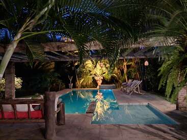 Esta imagem mostra uma área de piscina externa serena à noite, cercada por palmeiras exuberantes, móveis de madeira, espreguiçadeiras e iluminação suave e quente, criando tranquilidade.