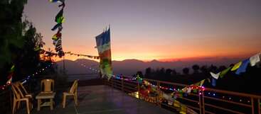 A serene terrace scene at dusk, adorned with colorful prayer flags and string lights, overlooking mountains. Three chairs and a table invite quiet contemplation.