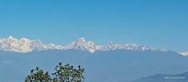 Snow-covered mountain peaks stand majestically under a clear blue sky. Green foliage is visible in the foreground. Crisp morning light enhances the landscape’s natural beauty and tranquility.