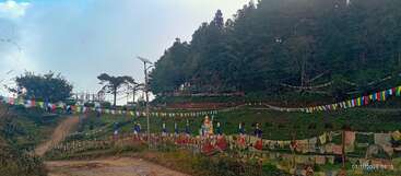 Colorful prayer flags flutter along a dirt road, surrounded by lush greenery, hills, and trees. A serene Buddhist stupa stands peacefully in the distance.