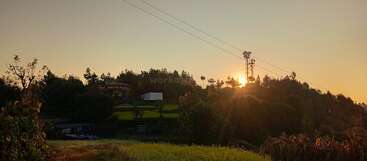 The image shows a serene rural landscape at sunset, with sunlight peeking through trees, lush green fields, a house, power lines, and a tall communication tower.