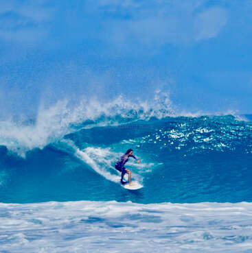 A surfer skillfully rides a large blue wave under a bright sky. Ocean spray surrounds them, showcasing excitement, athleticism, and the beauty of surfing adventures.