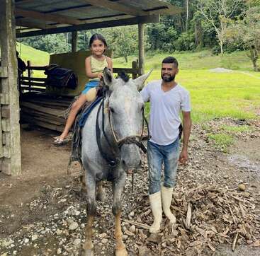 Une jeune fille souriante est assise sur un cheval gris, tandis qu'un homme, chaussé de bottes blanches, se tient à ses côtés. Ils sont en plein air, près d'une cabane en bois, entourés de verdure.