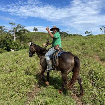 Un homme vêtu d'une chemise et d'un chapeau verts traverse un champ herbeux à cheval, en levant le pouce. Le ciel est bleu avec des nuages épars.