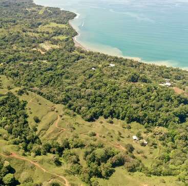 Cette image aérienne montre des forêts verdoyantes et des collines ondulantes rencontrant un littoral bleu clair, avec des maisons éparses et des chemins de terre sinueux à travers le paysage.