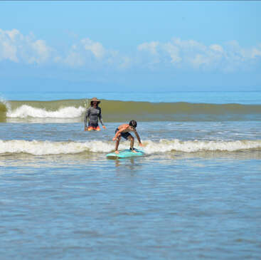 Un jeune garçon apprend à surfer sur de douces vagues tandis qu'un moniteur observe la scène. Le ciel est clair et bleu, créant une scène de plage paisible.