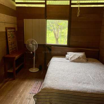 Cette chambre confortable en bois comprend un lit avec des draps rayés, un ventilateur sur pied, un petit bureau, un tableau encadré, une fenêtre avec vue sur l'extérieur et la lumière naturelle. Décor simple et rustique.