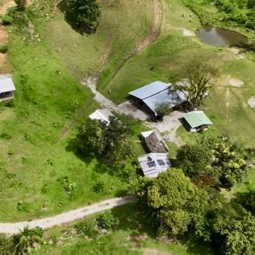 Vue aérienne d'une zone rurale avec des maisons dispersées, de l'herbe verte, des arbres, une route en gravier, un petit étang et une végétation luxuriante dans un cadre rural paisible.