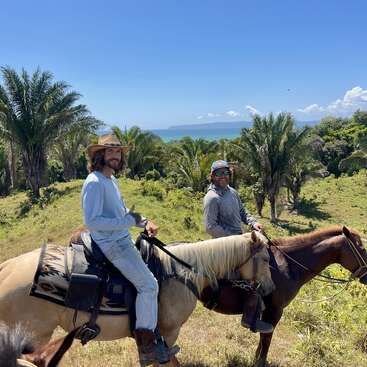 Deux hommes montent à cheval dans un paysage tropical luxuriant, peuplé de palmiers. Ils sourient sous un ciel d'un bleu éclatant, l'océan étant visible derrière eux.
