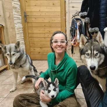 L'image montre un couple assis par terre avec deux huskies, un grand et un petit, dans une pièce confortable aux murs en bois et au sol carrelé.