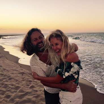 L'image représente un homme et une femme s'embrassant sur une plage de sable, avec l'océan et le coucher de soleil visibles en arrière-plan, transmettant un sentiment de bonheur et d'affection.