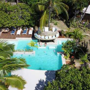 Une scène tropicale au bord d'une piscine, avec un bar aquatique, des palmiers, des chaises longues, une végétation luxuriante et des clients se prélassant au soleil dans un cadre extérieur somptueux.