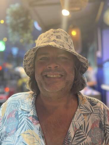 Un homme joyeux aux cheveux longs, coiffé d'un bob à motifs et vêtu d'une chemise aux couleurs tropicales, sourit dans un décor de soirée lumineux et animé, sans doute urbain.
